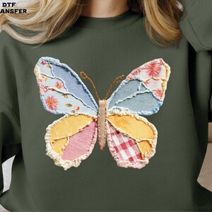 May include: Dark green sweatshirt featuring a colorful butterfly applique. The butterfly design incorporates various fabric patterns, including floral, denim, and gingham, in shades of blue, pink, yellow, and red. The text "DTF TRANSFER" is in the upper left corner.