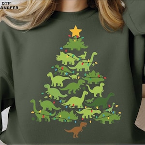 May include: Dark green sweatshirt featuring a Christmas tree design made of green dinosaurs, a yellow star, and string lights. A brown dinosaur is at the bottom. The text "DTF TRANSFER" is in the top left corner.