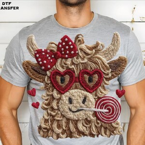 Pode incluir: Camiseta cinza claro com um design bordado extravagante de uma vaca das Terras Altas. A vaca tem &oacute;culos em forma de cora&ccedil;&atilde;o, um la&ccedil;o vermelho com bolinhas brancas e um pirulito. Pequenos cora&ccedil;&otilde;es vermelhos est&atilde;o espalhados pelo design.
