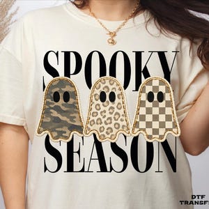 May include: A cream-colored t-shirt featuring the black text SPOOKY SEASON and three ghost illustrations. The ghosts have black oval eyes and are decorated with camouflage, leopard print, and a tan and brown checkerboard pattern respectively.