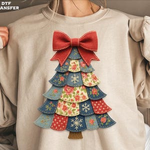 May include: A beige sweatshirt featuring a Christmas tree design. The tree is constructed from layered fabric in shades of blue, red, and floral patterns, topped with a red bow. Snowflake details are also present on the tree.