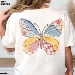 May include: Cream-colored t-shirt featuring a colorful butterfly applique on the back. The butterfly design incorporates various fabrics in shades of blue, yellow, pink, and red, with floral and gingham patterns. The text "DTF TRANSFER" is in the upper left corner.