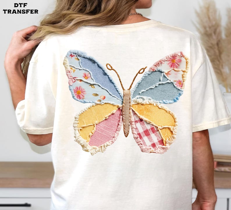 May include: Cream-colored t-shirt featuring a colorful butterfly applique on the back. The butterfly design incorporates various fabrics in shades of blue, yellow, pink, and red, with floral and gingham patterns. The text "DTF TRANSFER" is in the upper left corner.