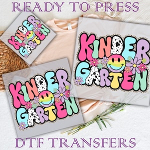 May include: Three iron-on transfers featuring the text "Kindergarten" in a colorful, bubbly font with a smiley face and flowers. The transfers are ready to press onto clothing or other items.