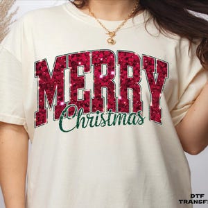 May include: A cream-colored short-sleeve t-shirt featuring the word MERRY in large, block-style, dark red glitter letters with a subtle sparkle effect. Below, the word Christmas is printed in a dark green, cursive script font.
