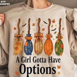 May include: Beige sweatshirt featuring five colorful broom designs with autumn leaves and bats. The text "A Girl Gotta Have Options" is printed in black. The brooms have different colors and patterns, with bows and patches.