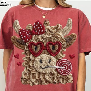 Pode incluir: Camiseta vermelha com um design bordado de uma vaca das Terras Altas com &oacute;culos de sol em forma de cora&ccedil;&atilde;o, um la&ccedil;o de bolinhas e um pirulito. A vaca tem um rosto sorridente e &eacute; cercada por cora&ccedil;&otilde;es bordados. O texto "DTF TRANSFER" est&aacute; no canto superior esquerdo.