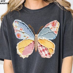 May include: Dark gray t-shirt featuring a colorful butterfly design. The butterfly wings are made of patchwork fabric in shades of blue, yellow, pink, and red, with floral and gingham patterns. The shirt has a relaxed fit.
