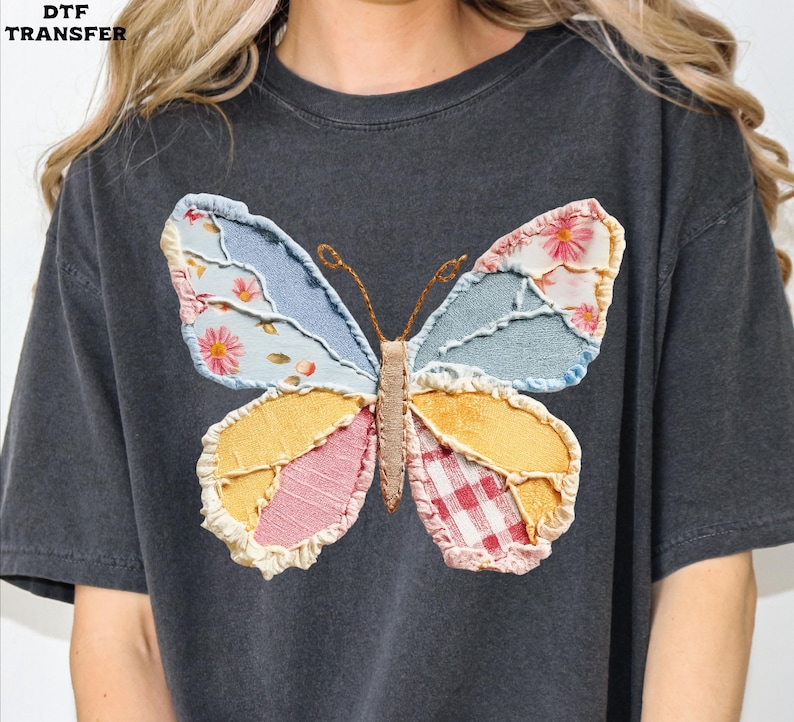 May include: Dark gray t-shirt featuring a colorful butterfly design. The butterfly wings are made of patchwork fabric in shades of blue, yellow, pink, and red, with floral and gingham patterns. The shirt has a relaxed fit.