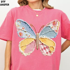 May include: Pink t-shirt featuring a large, patchwork butterfly design. The butterfly has wings in shades of blue, yellow, pink, and white, with floral and gingham patterns. The text "DTF TRANSFER" is in the upper left corner.