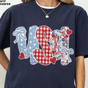 May include: Navy blue t-shirt with a patriotic "USA" design. The letters are appliqued with various patterns: blue stars, red gingham, and floral prints. Small red heart accents and bows complete the design.