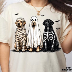 May include: A cream-colored t-shirt featuring a graphic of three dogs. The left dog is wrapped like a mummy, the center dog wears a white ghost sheet, and the right dog has a white skeleton print on its black fur. Small black bats fly above.