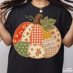 Peut inclure: Un t-shirt noir arborant un grand motif central de citrouille composé de divers empiècements en tissu. Le design de la citrouille présente des motifs floraux, à carreaux et à pois dans des tons orange, vert et beige, avec de la dentelle blanche.