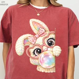 May include: Red t-shirt featuring a cartoon bunny with heart-shaped glasses and a pink bow. The bunny is blowing a bubblegum bubble. The shirt has the text "DTF TRANSFER" in the upper left corner.
