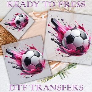 May include: A white soccer ball with black panels is surrounded by pink and black paint splatters. The image is on a white background and says "DTG Transfers" at the bottom.