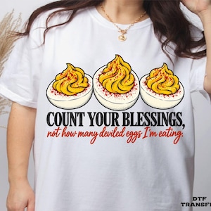 May include: A white t-shirt featuring a graphic of three halved deviled eggs with yellow yolks and red paprika seasoning. Below the eggs, the text reads: COUNT YOUR BLESSINGS, not how many deviled eggs I'm eating. The bottom right corner displays DTF TRANSFER.