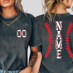 May include: Dark gray t-shirts with baseball-themed designs. One shirt features the number "00" in a baseball stitch design. The other shirt displays the word "NAME" in a baseball stitch design, with red stitching accents.