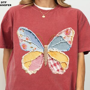 May include: A red-orange t-shirt featuring a large, colorful butterfly applique. The butterfly wings are patchwork, with floral, denim, and gingham patterns. The shirt has short sleeves and a crew neck.