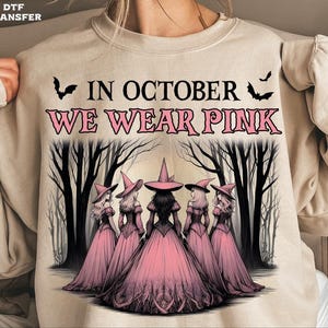 May include: Beige sweatshirt with a graphic that reads "IN OCTOBER WE WEAR PINK". The graphic features five witches in pink dresses and hats, with bats and a forest background. A Halloween-themed design.