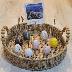 Puede incluir: Una cesta de mimbre con un asa de madera contiene una colección de huevos de cerámica coloridos. Cada huevo está unido a un clip metálico y un alambre delgado. Los huevos están decorados con varios patrones y colores, incluyendo rojo, azul, amarillo y blanco. La cesta está sentada sobre una superficie de madera.