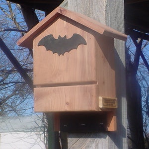 May include: A wooden bat house with a dark bat silhouette cutout on the front. The house is made of light brown wood and has a small rectangular sign on the lower right side. It is mounted on a wooden post.