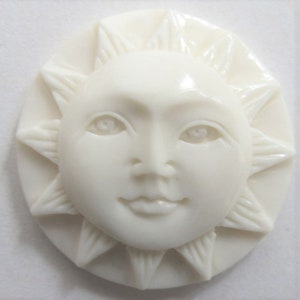 May include: A cream-colored sun-shaped cameo with a smiling face and radiating sun rays. The sun's face has detailed features, including eyes, a nose, and a smiling mouth. The cameo is oval-shaped.