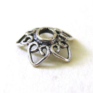 May include: Silver-toned metal bead cap with a decorative, openwork design resembling a flower. The bead cap has a round hole in the center.