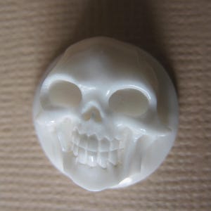 May include: A white carved skull made of bone. The skull has a detailed carving of the teeth and eye sockets.