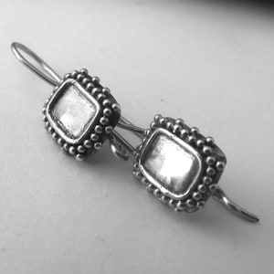 May include: A pair of silver earrings. Each earring has a square, recessed centre framed by a beaded edge. The earrings have a hook closure. The design is simple and elegant, suitable for everyday wear.