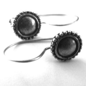 May include: A pair of silver-coloured earrings. Each earring has a circular, raised centre with a beaded border. The earrings have a simple, curved wire design. The earrings are shown against a white background.