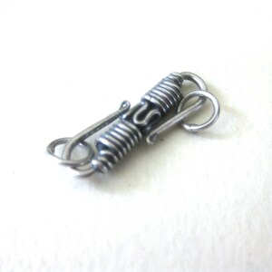 May include: A silver-toned metal clasp with a coiled design and a hook closure. The clasp features a decorative, spiraled center section and is designed for jewelry or craft projects.