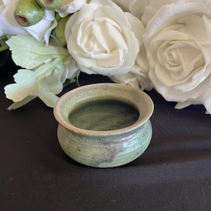 May include: A small, handcrafted ceramic bowl with a rounded shape and a wide opening. The bowl has a mottled green and tan glaze, with visible texture. The background includes white roses and a black surface.