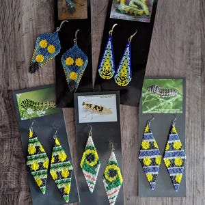 May include: A collection of beaded earrings in various geometric shapes and colors, including blue, yellow, green, and white. Some earrings feature insect imagery on the accompanying cards. The earrings are displayed on a wooden surface.