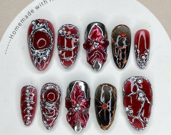 Puede incluir: Un conjunto de diez puntas de uñas artificiales de varios tamaños. Las uñas presentan una base rojo oscuro con detalles plateados y blancos. Algunas uñas tienen un diseño floral, otras un patrón de telaraña. El texto "Nail Tip" es visible.