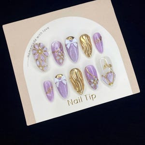 Puede incluir: Un conjunto de diez u&ntilde;as postizas de color lavanda con varios dise&ntilde;os, incluyendo detalles dorados, lazos y adornos florales. Las u&ntilde;as postizas est&aacute;n dispuestas sobre una tarjeta beige con la inscripci&oacute;n "Nail Tip" en letras doradas.