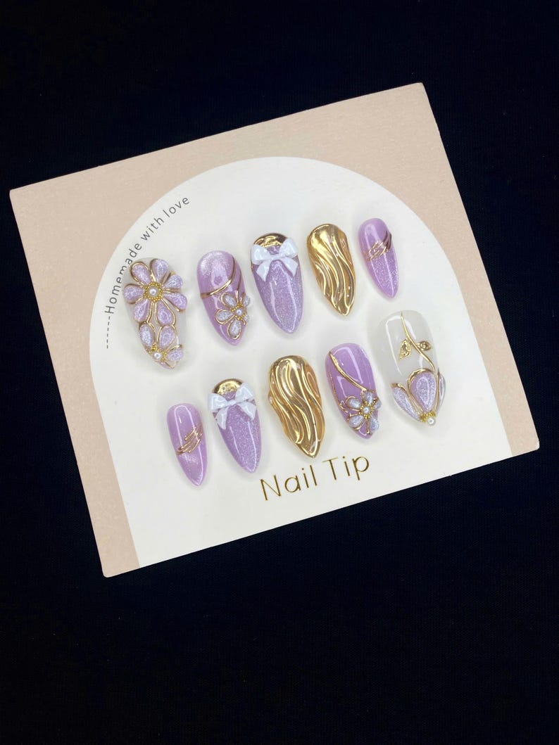 Puede incluir: Un conjunto de diez u&ntilde;as postizas de color lavanda con varios dise&ntilde;os, incluyendo detalles dorados, lazos y adornos florales. Las u&ntilde;as postizas est&aacute;n dispuestas sobre una tarjeta beige con la inscripci&oacute;n "Nail Tip" en letras doradas.