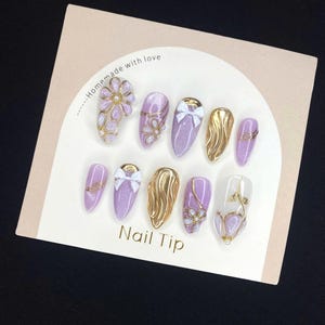 Puede incluir: Un conjunto de u&ntilde;as postizas en tonos lavanda y dorado con diversos dise&ntilde;os, incluyendo detalles florales, lazos y purpurina. El texto "Nail Tip" es visible en la tarjeta, junto con "Homemade with love".
