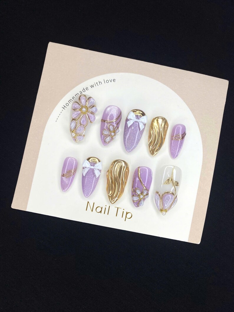 Puede incluir: Un conjunto de u&ntilde;as postizas en tonos lavanda y dorado con diversos dise&ntilde;os, incluyendo detalles florales, lazos y purpurina. El texto "Nail Tip" es visible en la tarjeta, junto con "Homemade with love".
