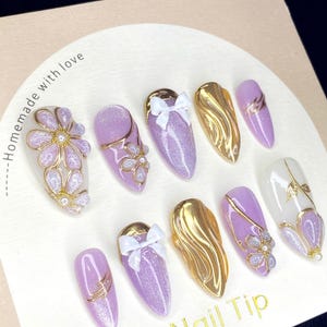 Puede incluir: Una colecci&oacute;n de diez u&ntilde;as artificiales con varios dise&ntilde;os. Las u&ntilde;as presentan una base de purpurina morada clara, detalles dorados y lazos blancos. Algunas u&ntilde;as tienen adornos florales y de perlas. El texto "Homemade with love" y "Nail Tip" son visibles.