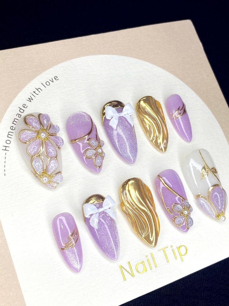 Puede incluir: Una colecci&oacute;n de diez u&ntilde;as artificiales con varios dise&ntilde;os. Las u&ntilde;as presentan una base de purpurina morada clara, detalles dorados y lazos blancos. Algunas u&ntilde;as tienen adornos florales y de perlas. El texto "Homemade with love" y "Nail Tip" son visibles.