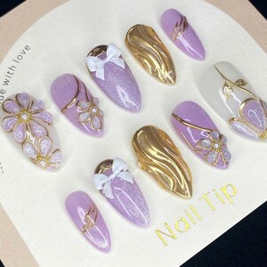 Puede incluir: Una colecci&oacute;n de puntas de u&ntilde;as artificiales en varios dise&ntilde;os. El conjunto incluye u&ntilde;as de purpurina lavanda con lazos blancos, u&ntilde;as met&aacute;licas doradas y u&ntilde;as con detalles florales y dorados. El texto "Nail Tip" est&aacute; impreso.