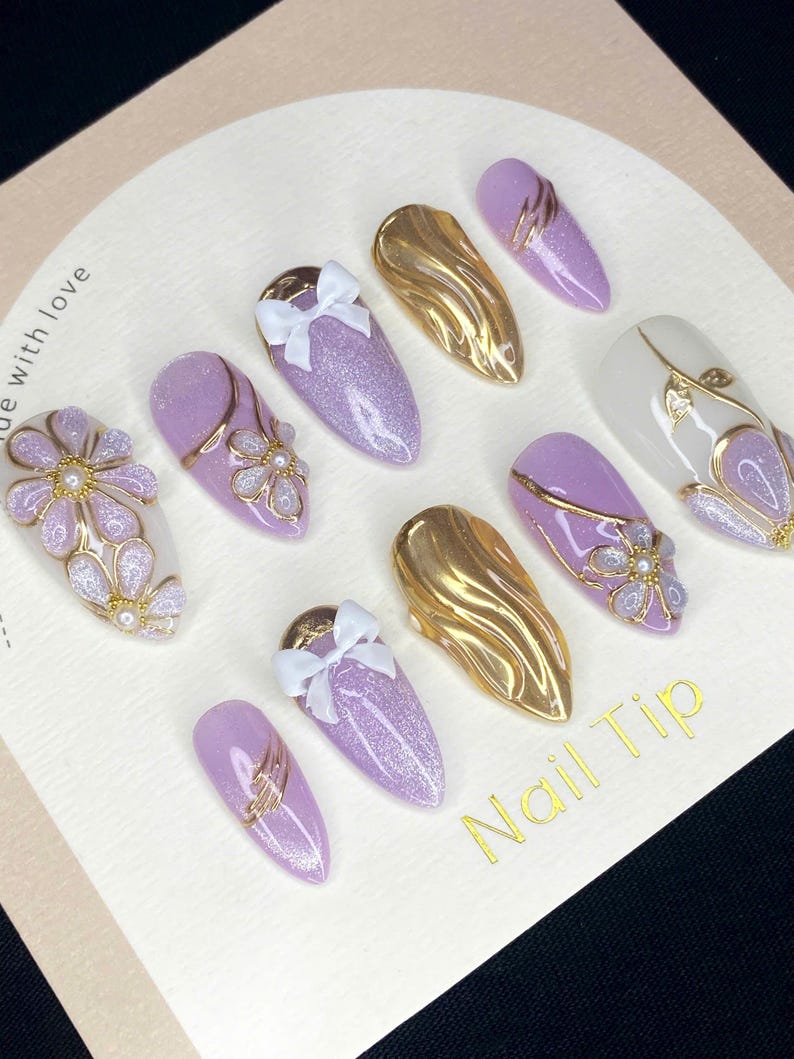 Puede incluir: Una colecci&oacute;n de puntas de u&ntilde;as artificiales en varios dise&ntilde;os. El conjunto incluye u&ntilde;as de purpurina lavanda con lazos blancos, u&ntilde;as met&aacute;licas doradas y u&ntilde;as con detalles florales y dorados. El texto "Nail Tip" est&aacute; impreso.