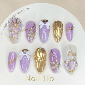 Puede incluir: Una colecci&oacute;n de diez u&ntilde;as postizas con varios dise&ntilde;os. Algunas son lavanda con purpurina, adornadas con detalles dorados, lazos blancos y adornos florales. Otras son doradas o blancas con detalles dorados. El texto "Nail Tip" est&aacute; impreso.