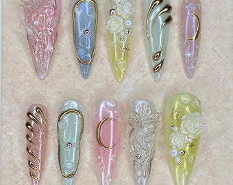 Op de afbeelding: Een verzameling van tien lange, puntige kunstnagels met gedetailleerde ontwerpen. De nagels hebben verschillende kleuren, waaronder roze, blauw en geel, met gouden accenten, bloemmotieven en sprankelende details. De tekst "Nail Art" staat bovenaan.