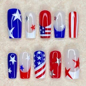 Pode incluir: Um conjunto de dez unhas artificiais com tema patriótico. Os designs incluem cores azul, vermelho e branco com padrões de estrelas e listras. O texto "Nail Art" é visível no topo.