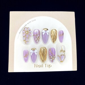 Puede incluir: Un juego de diez u&ntilde;as artificiales con varios dise&ntilde;os. Las u&ntilde;as presentan una mezcla de purpurina lavanda, dorado y detalles transparentes. Algunas u&ntilde;as tienen lazos dorados, dise&ntilde;os florales o patrones de ondas doradas. La tarjeta dice "Homemade with love" y "Nail Tip".