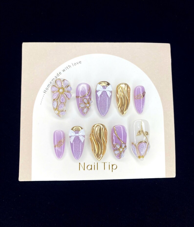 Puede incluir: Un juego de diez u&ntilde;as artificiales con varios dise&ntilde;os. Las u&ntilde;as presentan una mezcla de purpurina lavanda, dorado y detalles transparentes. Algunas u&ntilde;as tienen lazos dorados, dise&ntilde;os florales o patrones de ondas doradas. La tarjeta dice "Homemade with love" y "Nail Tip".