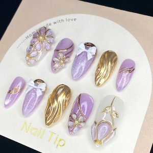 Puede incluir: Una colecci&oacute;n de u&ntilde;as postizas con diversos dise&ntilde;os. Las u&ntilde;as presentan colores dorados, lavanda y blanco, adornadas con lazos, detalles florales y purpurina. El texto "Nail Tip" es visible.