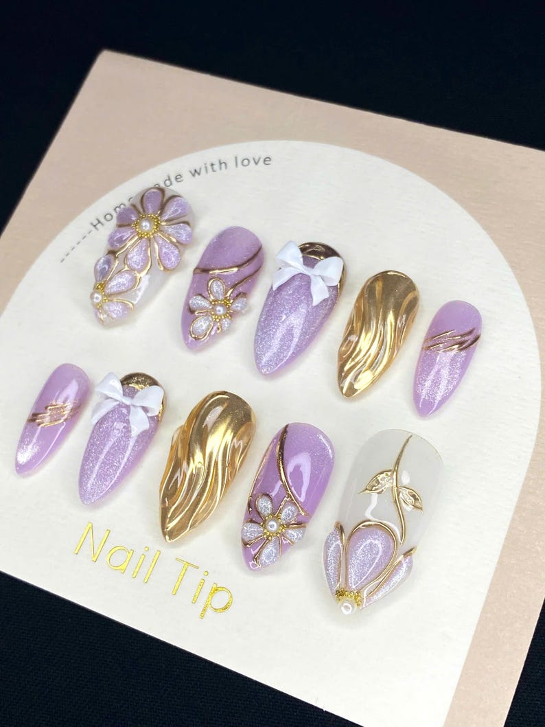 Puede incluir: Una colecci&oacute;n de u&ntilde;as postizas con diversos dise&ntilde;os. Las u&ntilde;as presentan colores dorados, lavanda y blanco, adornadas con lazos, detalles florales y purpurina. El texto "Nail Tip" es visible.