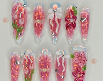 Può includere: Un set di dieci unghie finte lunghe a forma di mandorla. Ogni unghia presenta un design rosa e iridescente con vari abbellimenti, tra cui fiocchi, fiori e strass. Il testo "Nailbox FOR YOU" è in alto e "Thank you for choosing us" in basso.